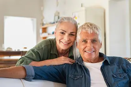 Portrait of happy senior couple relaxing at home and looking at camera. Romantic mature woman embrace from behind her smiling old husband. Cheerful senior couple enjoying life and retirement.