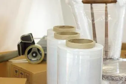 Approach to plastic rolls for packing in the foreground with a plastic wrapped chair, cardboard boxes and adhesive tape in the background