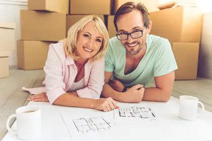 Beautiful mature couple in casual clothes is discussing plan of their new house, looking at camera and smiling while lying on the floor near boxes for move